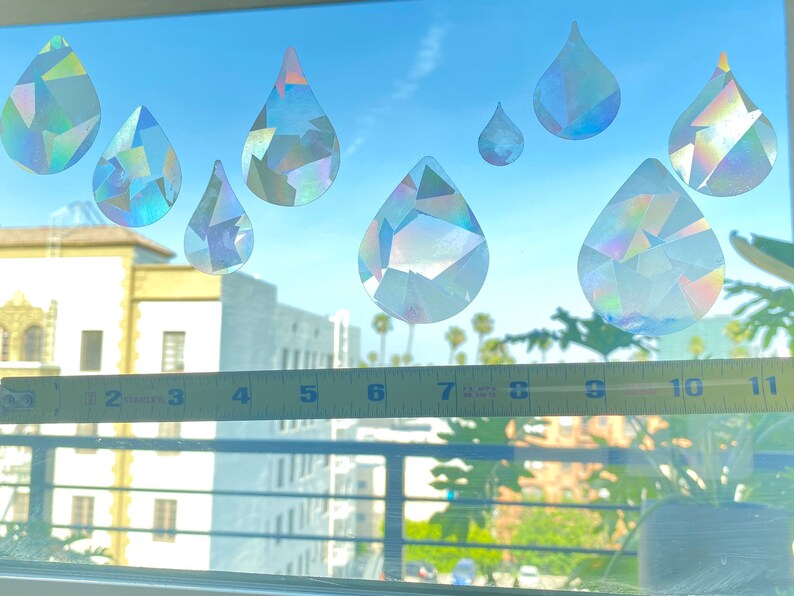 May include: A window with seven iridescent rainbow-colored raindrop stickers on it. A yellow measuring tape is placed on the window sill.