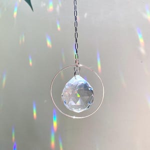 May include: A clear crystal ball hanging from a silver chain within a silver ring. The crystal ball is faceted and casts rainbows on the wall behind it.