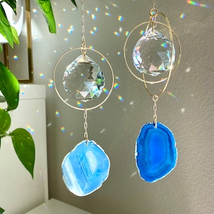 May include: Two crystal suncatchers with agate slices hanging from gold chains. The crystal suncatchers are clear and have a faceted design. The agate slices are blue and light blue.