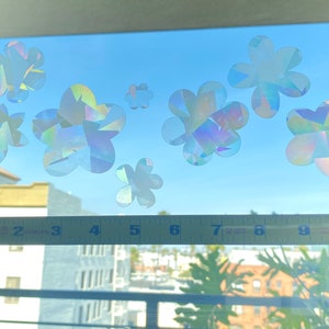 May include: A window with a blue sky and a cityscape in the background. There are several iridescent flower stickers on the window. The stickers are in various sizes and shapes. A yellow measuring tape is placed across the bottom of the window.