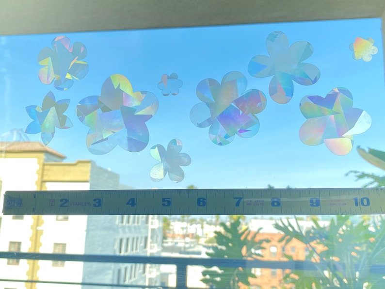 May include: A window with a blue sky and a cityscape in the background. There are several iridescent flower stickers on the window. The stickers are in various sizes and shapes. A yellow measuring tape is placed across the bottom of the window.