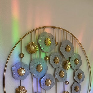 May include: A gold metal wall decor with a circular frame and multiple gray metal flowers with gold centers. The flowers are arranged in a circular pattern and are attached to the frame with gold metal rods.