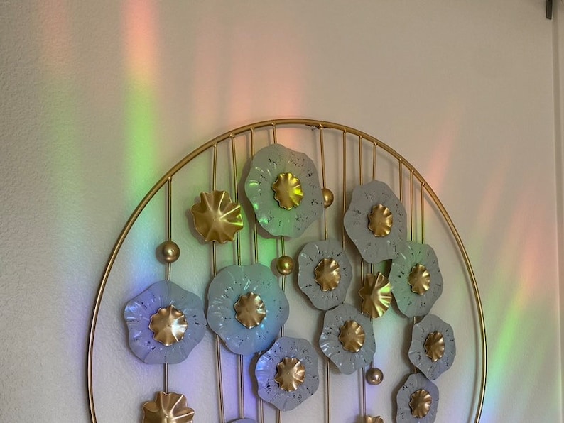 May include: A gold metal wall decor with a circular frame and multiple gray metal flowers with gold centers. The flowers are arranged in a circular pattern and are attached to the frame with gold metal rods.
