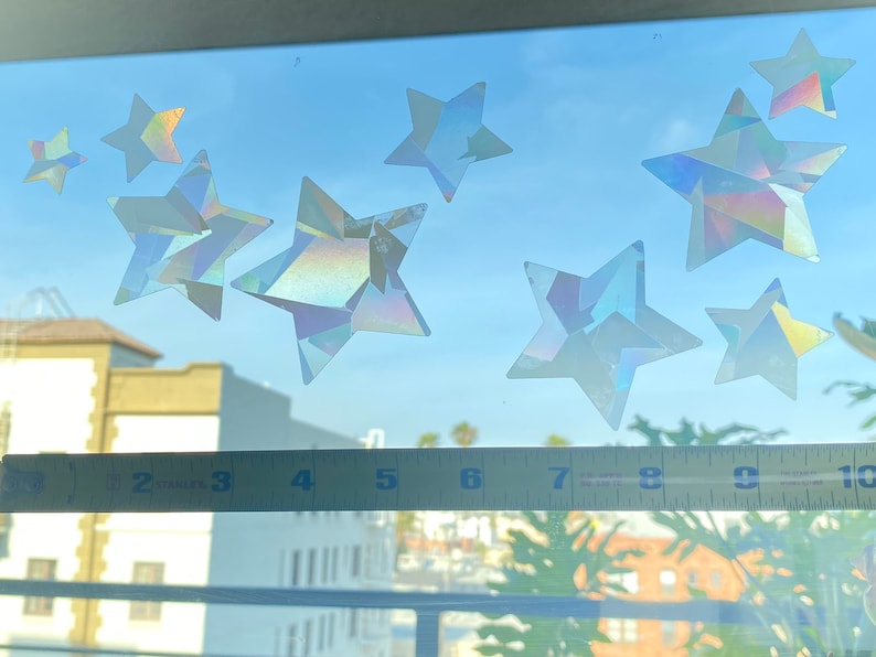 May include: A window with a blue sky and white clouds. There are nine iridescent star stickers on the window. A yellow ruler is placed on the windowsill.