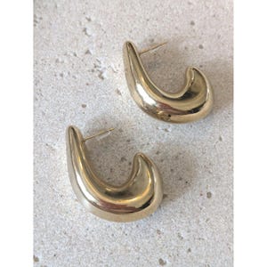 May include: A pair of gold-toned earrings with a smooth, curved, teardrop-like shape. Each earring features a polished, reflective surface and a thin metal post with a clear plastic backing, designed to be worn as a stud-style accessory.