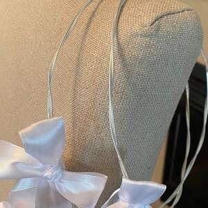 May include: A silver wire headpiece with multiple white satin bows. The bows are evenly spaced along the wire, creating a delicate and elegant design. The headpiece is displayed on a mannequin head.