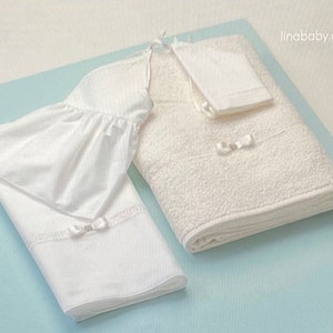 May include: A white baby blanket set with a ruffled edge, a small white towel with a bow, and a larger white towel with a bow. The towels and blanket are on a light blue surface.