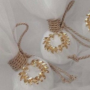 May include: Two white, round, fabric ornaments with gold leaf wreaths. The ornaments are tied with beige ribbon and are on a white tulle background.