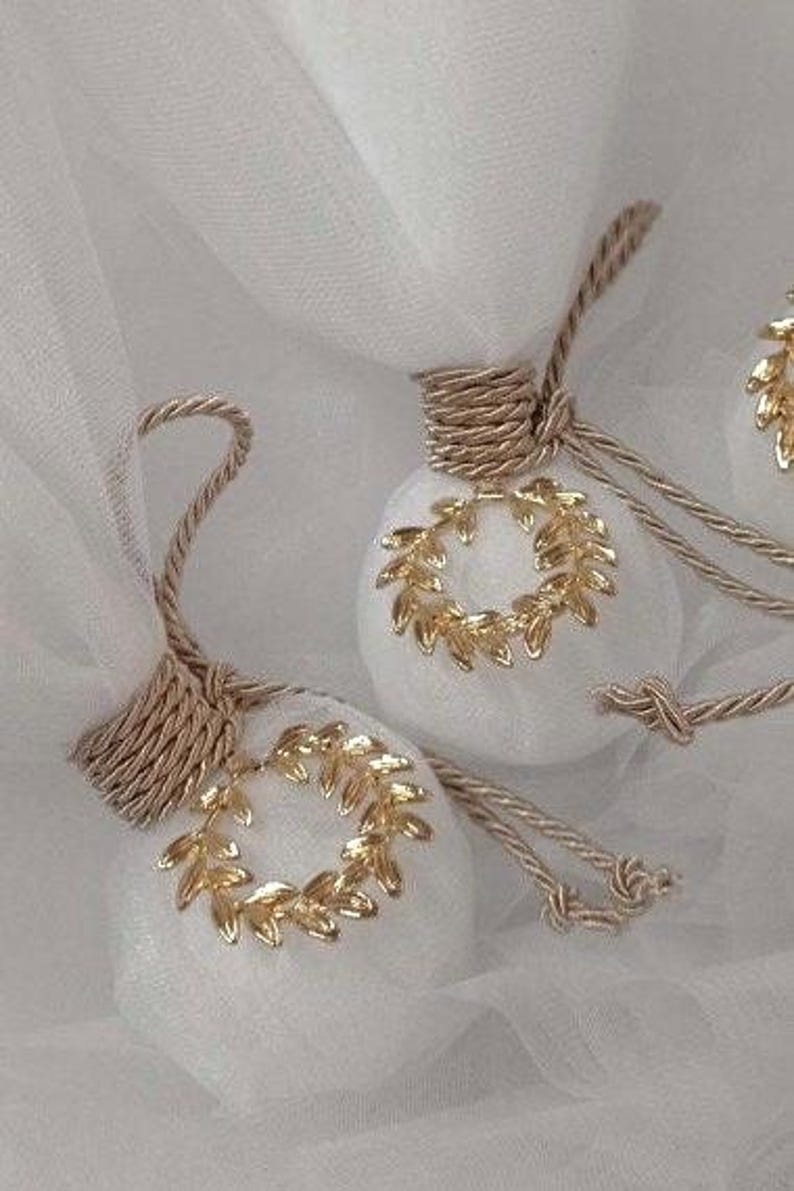 May include: Two white, round, fabric ornaments with gold leaf wreaths. The ornaments are tied with beige ribbon and are on a white tulle background.
