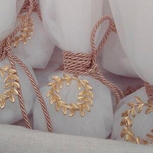 May include: A collection of small white fabric bags tied with pink rope and decorated with gold leaf wreaths. The bags are arranged on a white fabric surface.