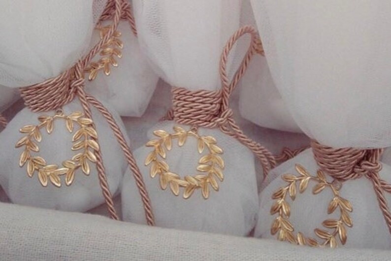May include: A collection of small white fabric bags tied with pink rope and decorated with gold leaf wreaths. The bags are arranged on a white fabric surface.