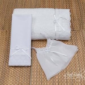 May include: A white christening gown with lace trim and a white satin ribbon bow, a white towel with lace trim and a white satin ribbon bow, and a white towel with lace trim.