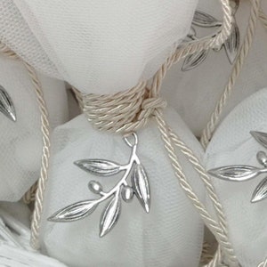May include: White tulle favor bags tied with ivory satin ribbon and adorned with silver olive branch charms. The bags are arranged in a white wicker basket.