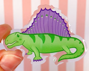 May include: A clear acrylic pin featuring a green Dimetrodon with dark green stripes and a prominent purple sail with small green dots along the top edge. The prehistoric reptile is depicted in profile with a visible mouth and four feet.
