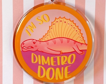 Könnte beinhalten: Ein runder Acryl-Schlüsselanhänger mit einer Illustration eines rosa Dimetrodons mit gelbem Segel. Das Design enthält den Text "I'M SO DIMETRO DONE" in gelber Schrift auf rosa Hintergrund, umrandet von einem orangefarbenen Rand und einem silbernen Metallverschluss.