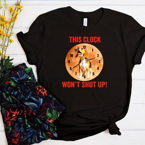 May include: Black t-shirt with a humorous graphic. The design features a beagle dog inside a clock face with the text "THIS CLOCK WON'T SHUT UP!" in red. The clock hands point to 12:00. The shirt is made of a soft material.