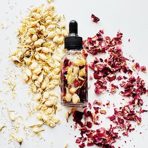 May include: A clear glass dropper bottle filled with a liquid and flower petals, with a black cap. Flanking the bottle are piles of dried jasmine buds and red rose petals, all on a white background. The image is a top-down view.