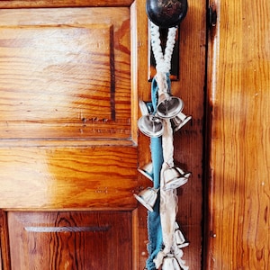 May include: A decorative door knocker featuring a series of small silver bells hanging from a braided rope and fabric. The bells are attached to a wooden door with a dark metal doorknob. The door has a warm, natural wood finish.