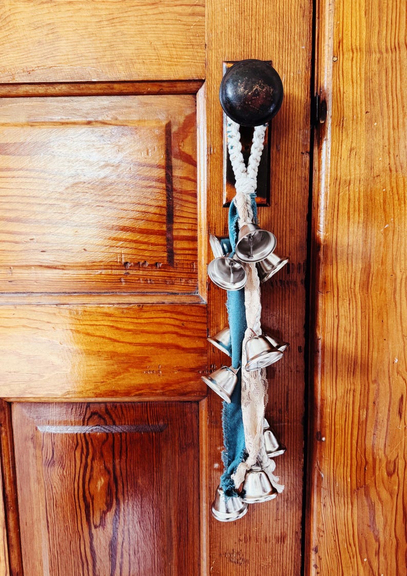 May include: A decorative door knocker featuring a series of small silver bells hanging from a braided rope and fabric. The bells are attached to a wooden door with a dark metal doorknob. The door has a warm, natural wood finish.