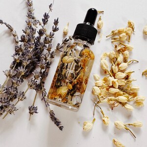 May include: A clear glass dropper bottle filled with a golden liquid and dried flowers, next to sprigs of lavender and jasmine blossoms. The bottle has a black cap and is surrounded by white petals.
