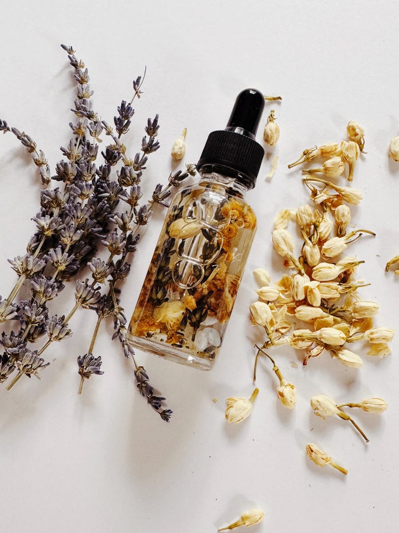 May include: A clear glass dropper bottle filled with a golden liquid and dried flowers, next to sprigs of lavender and jasmine blossoms. The bottle has a black cap and is surrounded by white petals.