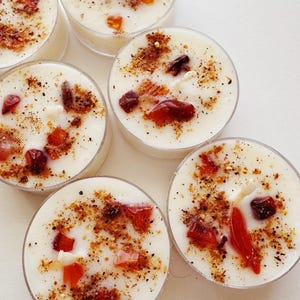 May include: A collection of six small, round candles in clear plastic containers. The white wax is speckled with dark flecks and adorned with red and orange crystals. These decorative candles are ideal for creating a relaxing ambiance.