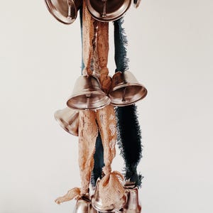 May include: A cluster of silver bells hangs from a collection of ribbons. The ribbons are in shades of tan, teal, and dark green. The bells are arranged in a decorative pattern, creating a visually appealing display.