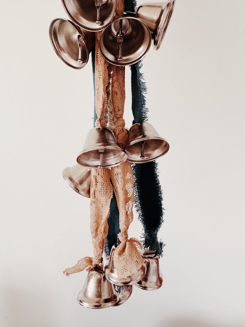 May include: A cluster of silver bells hangs from a collection of ribbons. The ribbons are in shades of tan, teal, and dark green. The bells are arranged in a decorative pattern, creating a visually appealing display.