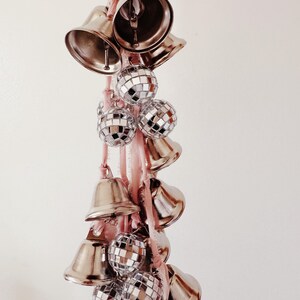 May include: A pink ribbon with silver bells and disco balls hanging from it.
