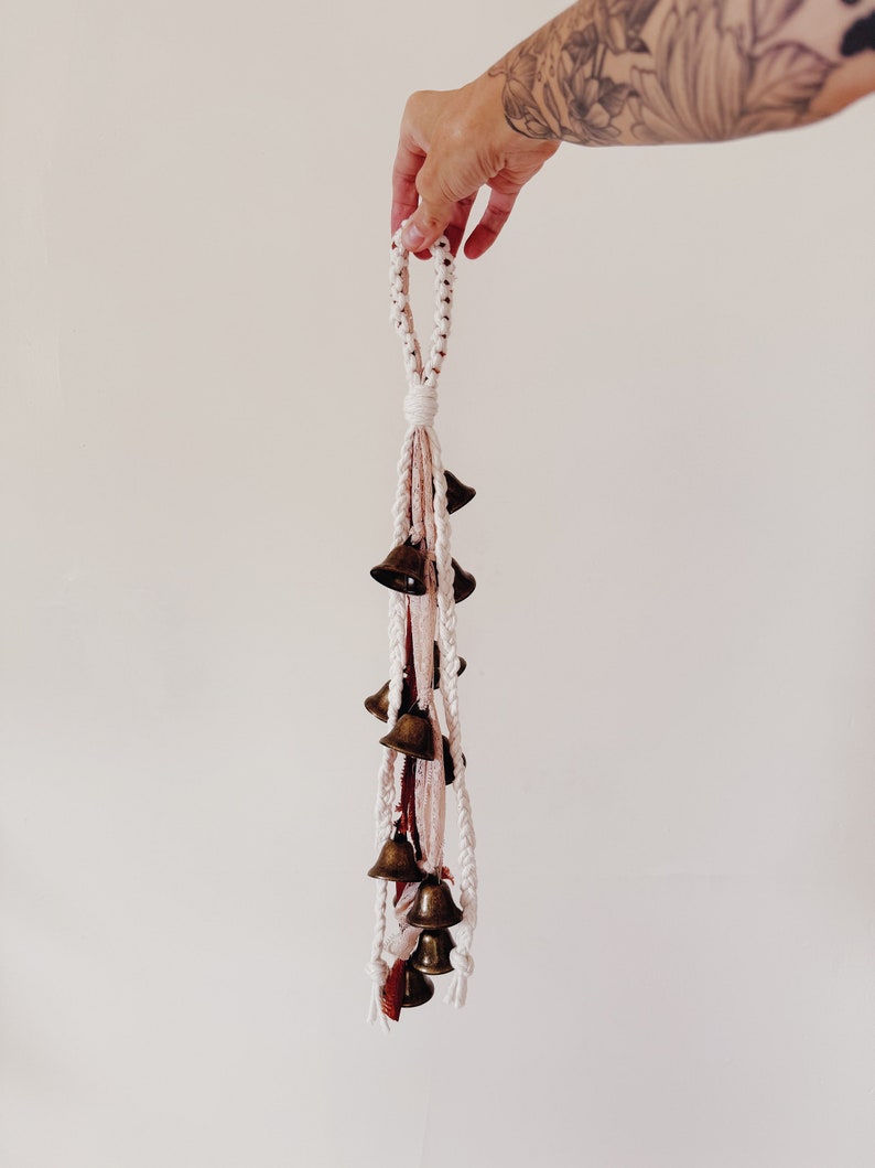 May include: A white macrame tassel with a loop at the top and a cluster of small brass bells hanging from the bottom.