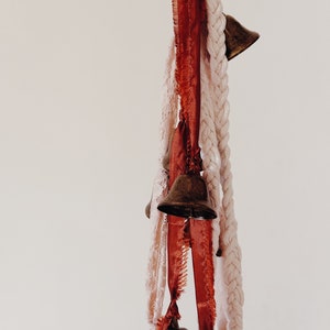 May include: A string of small brass bells hangs from a white and red ribbon. The bells are tied together with a white braided rope.