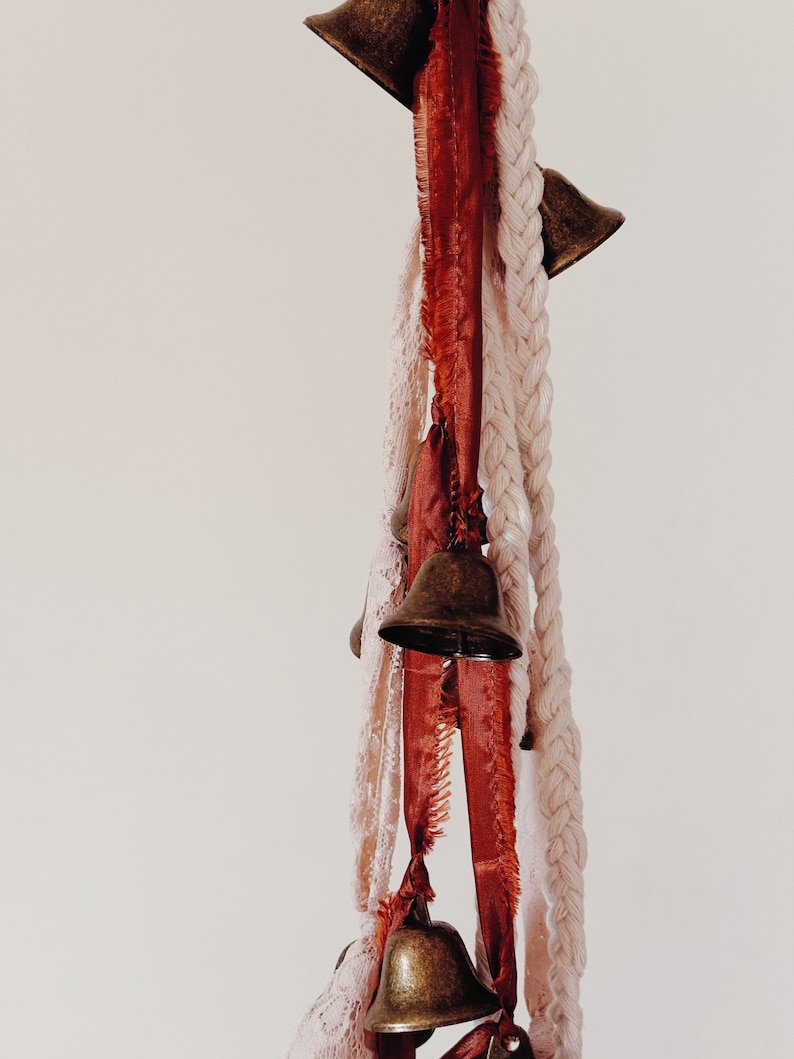 May include: A string of small brass bells hangs from a white and red ribbon. The bells are tied together with a white braided rope.