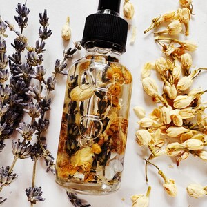 May include: A clear glass bottle with a black dropper filled with dried flowers and herbs. The bottle is surrounded by sprigs of lavender and dried jasmine flowers, creating a natural and botanical aesthetic. The product is likely a beauty or wellness oil.