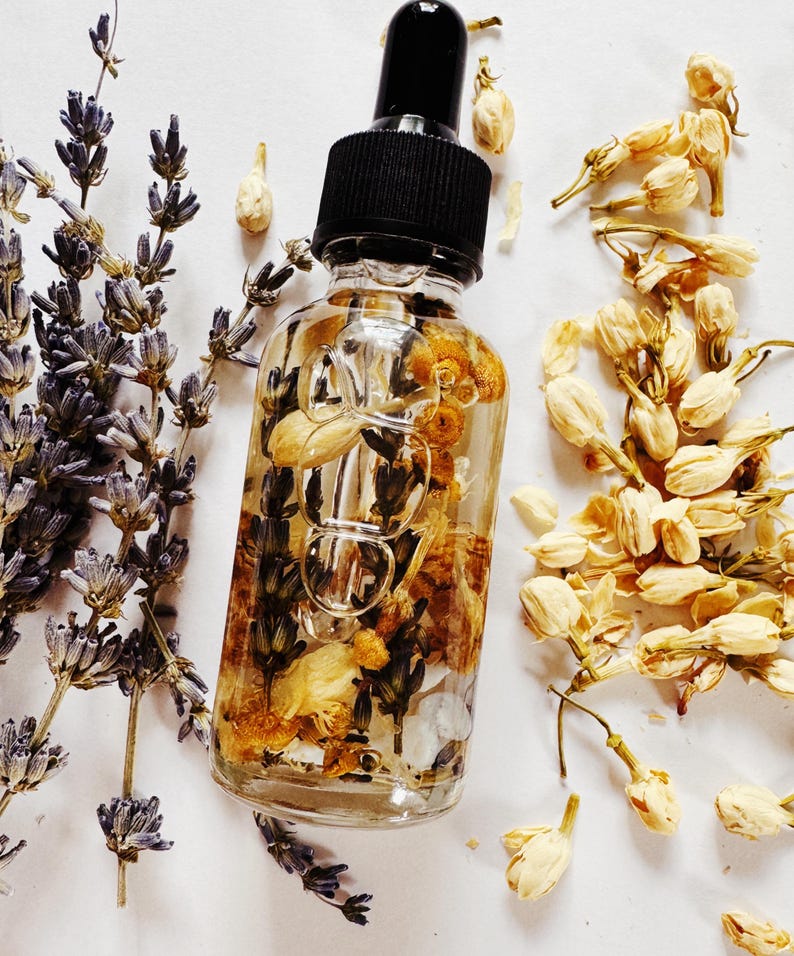 May include: A clear glass bottle with a black dropper filled with dried flowers and herbs. The bottle is surrounded by sprigs of lavender and dried jasmine flowers, creating a natural and botanical aesthetic. The product is likely a beauty or wellness oil.