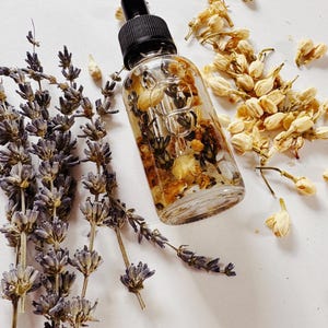 May include: A clear glass dropper bottle with a black cap, filled with dried flowers and herbs. Lavender sprigs and dried jasmine flowers surround the bottle, creating a natural, botanical aesthetic. The product is likely a skincare oil or serum.