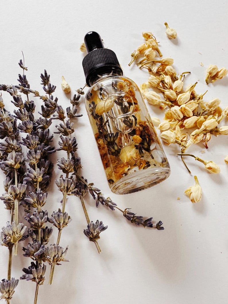 May include: A clear glass dropper bottle with a black cap, filled with dried flowers and herbs. Lavender sprigs and dried jasmine flowers surround the bottle, creating a natural, botanical aesthetic. The product is likely a skincare oil or serum.