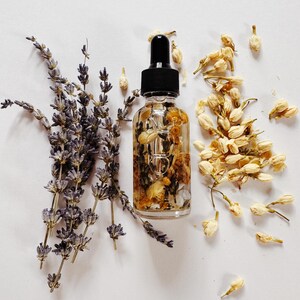 May include: A clear glass dropper bottle with a black cap, filled with oil and dried flowers. Lavender sprigs and dried jasmine buds surround the bottle, creating a natural, botanical aesthetic. The image is set against a white background.