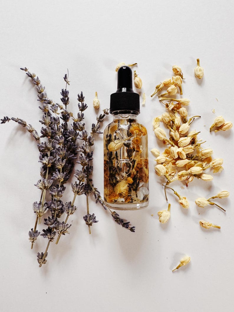 May include: A clear glass dropper bottle with a black cap, filled with oil and dried flowers. Lavender sprigs and dried jasmine buds surround the bottle, creating a natural, botanical aesthetic. The image is set against a white background.