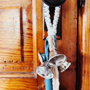 May include: A decorative door knocker featuring a dark metal knob and a braided rope with silver bells and teal fabric accents. The bells are attached to a wooden door.