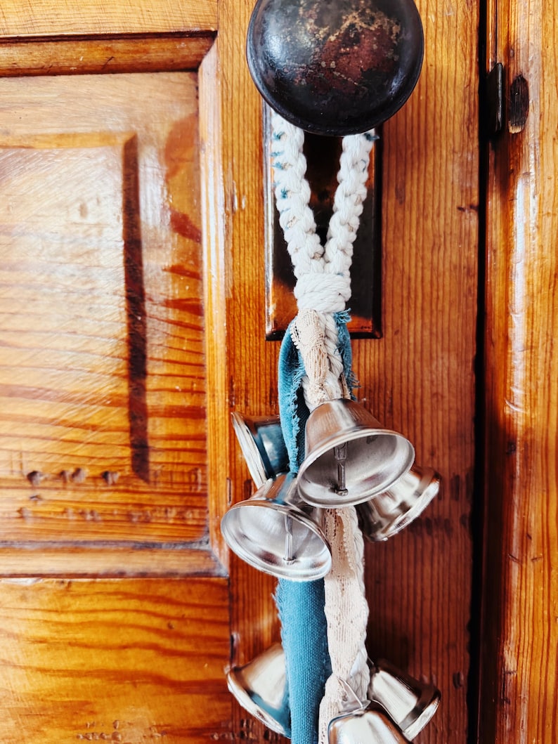 May include: A decorative door knocker featuring a dark metal knob and a braided rope with silver bells and teal fabric accents. The bells are attached to a wooden door.