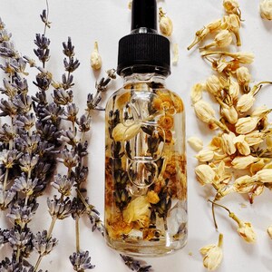 May include: A clear glass dropper bottle filled with dried flowers and herbs, topped with a black cap. Lavender sprigs and dried jasmine flowers surround the bottle. The bottle contains yellow and brown dried flowers.
