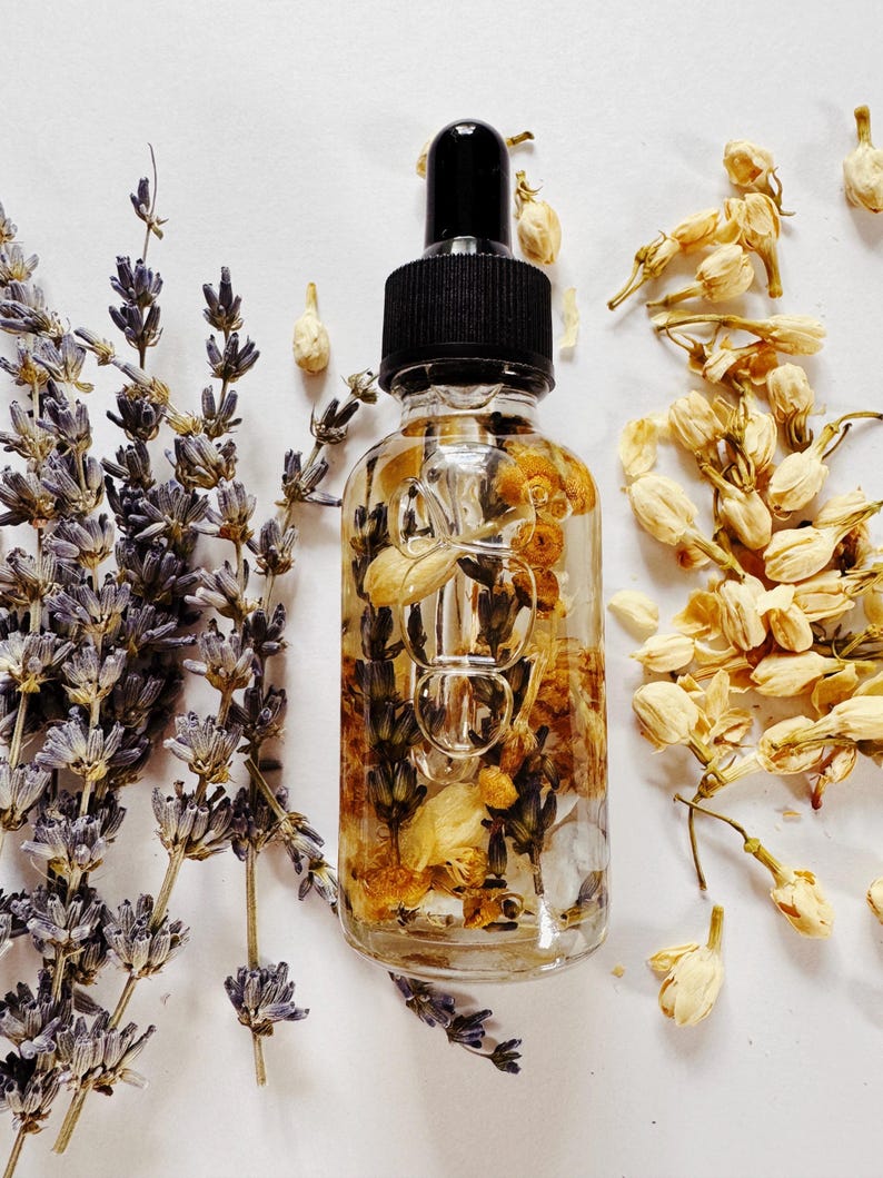 May include: A clear glass dropper bottle filled with dried flowers and herbs, topped with a black cap. Lavender sprigs and dried jasmine flowers surround the bottle. The bottle contains yellow and brown dried flowers.