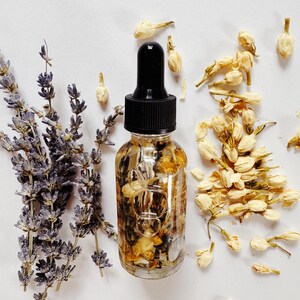 May include: A clear glass dropper bottle filled with dried flowers and herbs, topped with a black cap. Lavender sprigs and dried jasmine flowers are arranged around the bottle on a white surface. The bottle contains a floral oil.