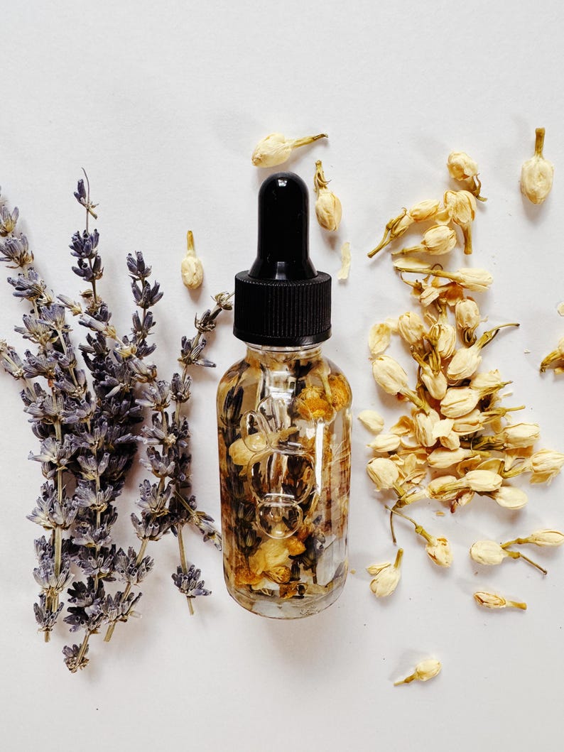 May include: A clear glass dropper bottle filled with dried flowers and herbs, topped with a black cap. Lavender sprigs and dried jasmine flowers are arranged around the bottle on a white surface. The bottle contains a floral oil.