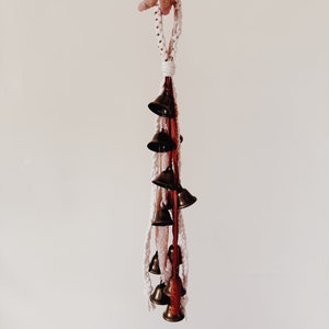 May include: A white and red macrame string with a loop at the top and a series of small brass bells hanging from the bottom.