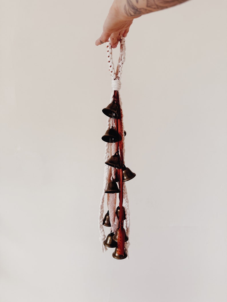 May include: A white and red macrame string with a loop at the top and a series of small brass bells hanging from the bottom.