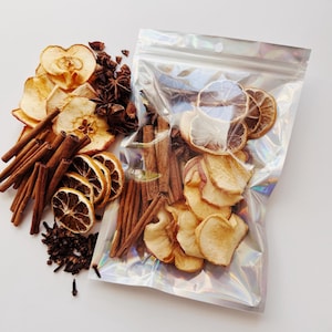 May include: A clear, iridescent bag filled with dried apple slices, orange slices, cinnamon sticks, star anise, and cloves. The ingredients are also arranged outside the bag on a white surface, creating a visually appealing display of natural elements.