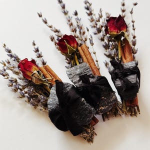 May include: Three bundles of dried lavender, a single dried red rose, and a cinnamon stick, each secured with a piece of black charcoal and tied together with a black ribbon. The bundles are arranged in a cluster against a plain white background.