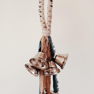 May include: A cluster of silver bells hangs from a braided rope handle. The rope is cream-colored with dark flecks. Below the rope are strips of blue and tan fabric. The bells are shiny and metallic.
