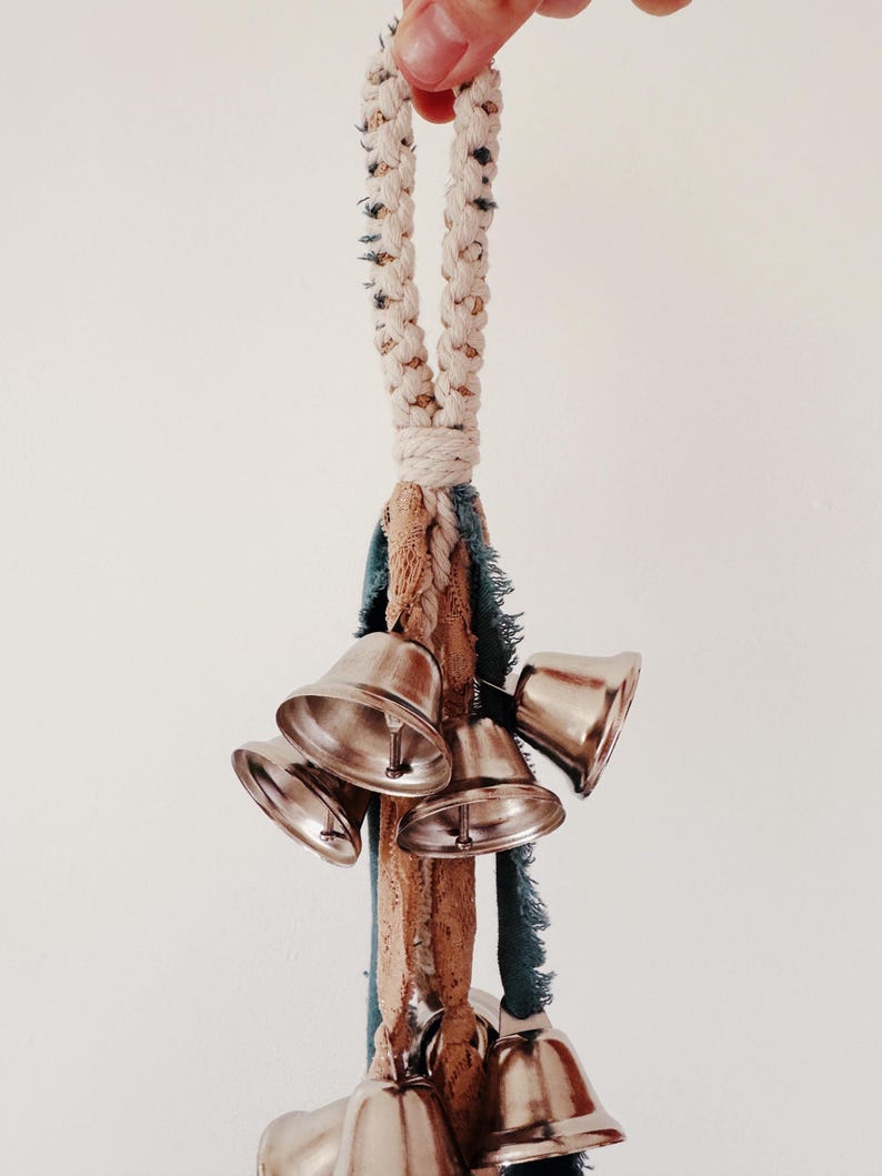 May include: A cluster of silver bells hangs from a braided rope handle. The rope is cream-colored with dark flecks. Below the rope are strips of blue and tan fabric. The bells are shiny and metallic.